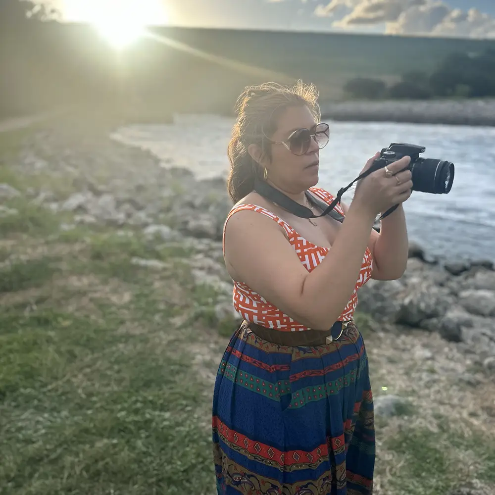 A woman holding and looking at a DSLR camera, surrounded by water and hills and greenery