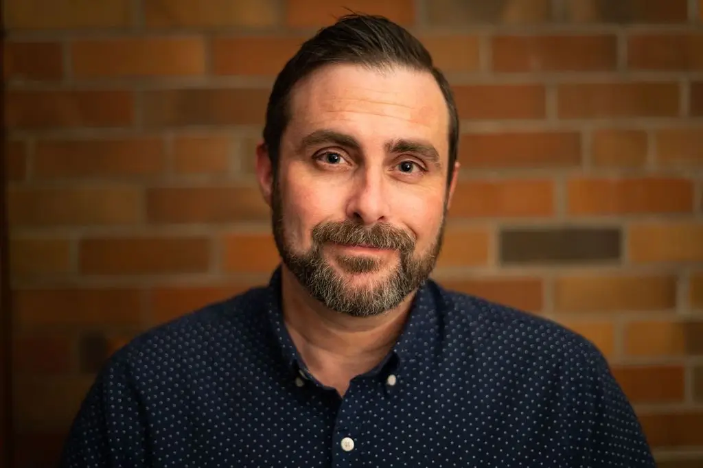 Headshot photo of a man in a blue shirt with a brick background behind him